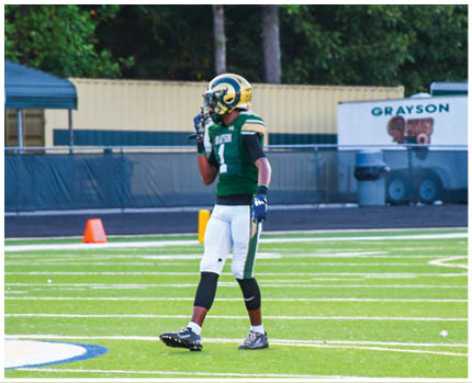 A person wearing a white and gold football uniform with the number 20 stands on a field while two other people in red and gold football uniforms are visible in the background near empty bleachers. Description generated by AI