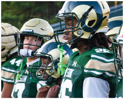 Three people wearing white and gold football uniforms and helmets are standing and interacting near a chain-link fence with trees and bleachers in the background. Description generated by AI