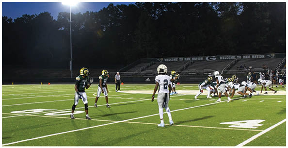 Four people wearing white football uniforms with gold helmets and large numbers on their backs are standing on a field. Description generated by AI