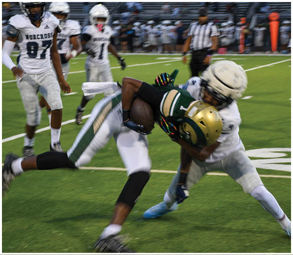 A person in a white football uniform with the number 12 and a gold helmet stands on a football field with blurred spectators and cheerleaders in the background. Description generated by AI