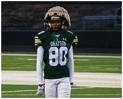 A person wearing a white football uniform with a gold and black helmet is kneeling on a green football field with arms extended forward. Description generated by AI