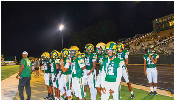 Four people wearing football helmets and uniforms are lined up facing an opponent on a field. Description generated by AI