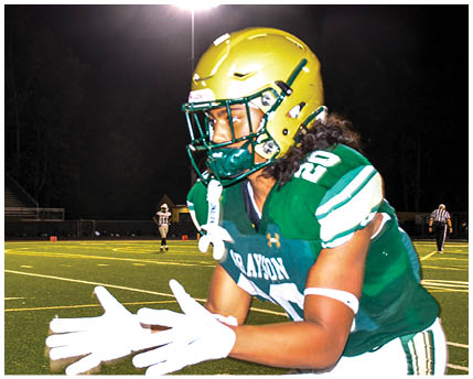 A person wearing sunglasses and a headset is speaking intensely while another person in a football helmet looks on in the background. Description generated by AI
