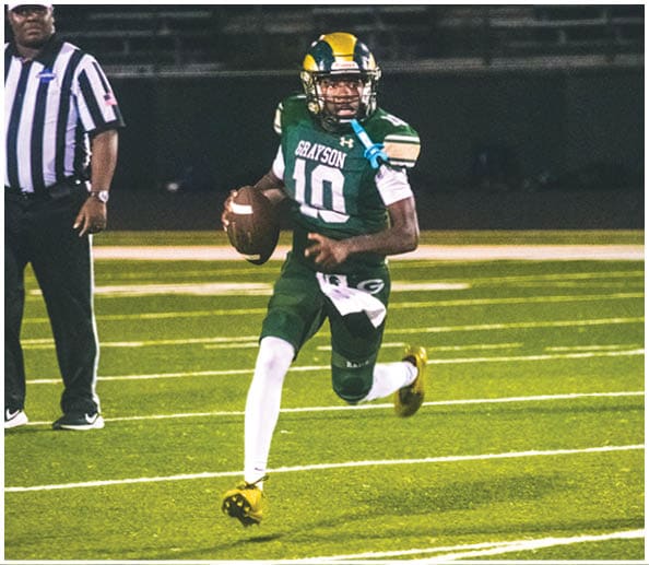 A person in a white and gold football uniform is crouched in an athletic stance on a green football field with other players and a coach in the background. Description generated by AI