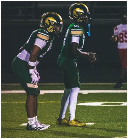 A person with long dreadlocks wearing a white football jersey with the name Grayson and the number 28 stands in front of two other people in similar football uniforms and helmets. Description generated by AI