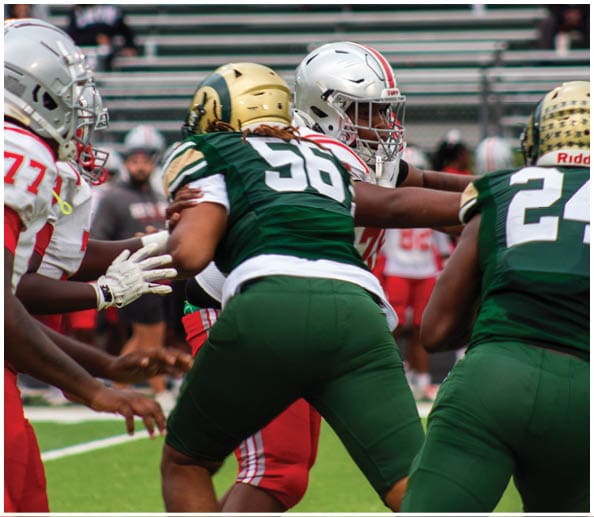 A person in a white and green football uniform with a gold helmet is on a field facing away from the camera while another person in a maroon and gold uniform stands in the background near empty bleachers. Description generated by AI
