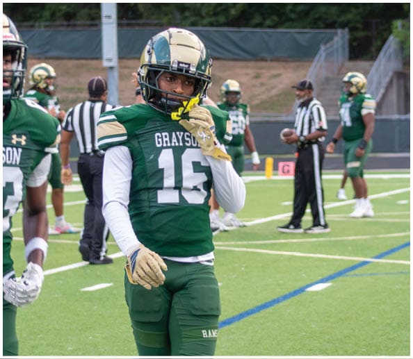 A person in a white and gold football uniform is sitting on the field holding a football while other players stand in the background. Description generated by AI