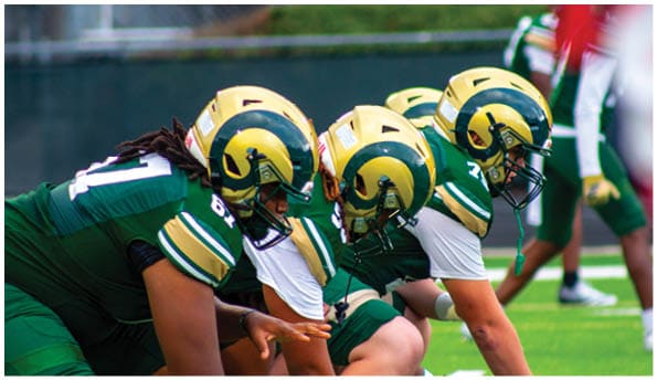 A person wearing a white football uniform and helmet is running with a football on a field while people in green cheerleading outfits stand in the background. Description generated by AI