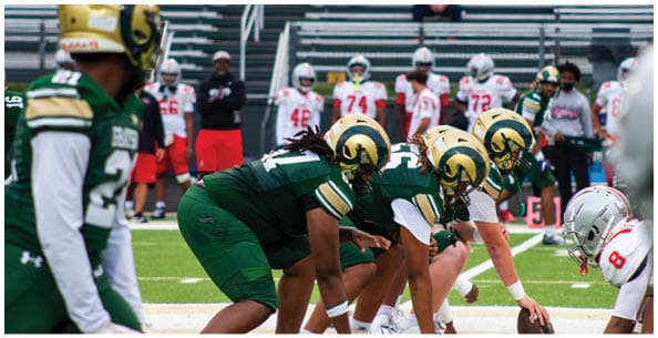 Four people wearing white football uniforms with gold helmets and large numbers on their backs are standing on a field. Description generated by AI