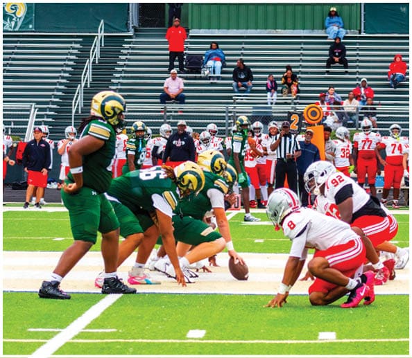 A person in a white football uniform with the number 12 and a gold helmet stands on a football field with blurred spectators and cheerleaders in the background. Description generated by AI