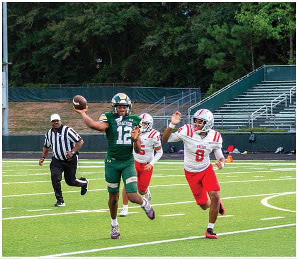A person in a white and gold football uniform is crouched in an athletic stance on a green football field with other players and a coach in the background. Description generated by AI