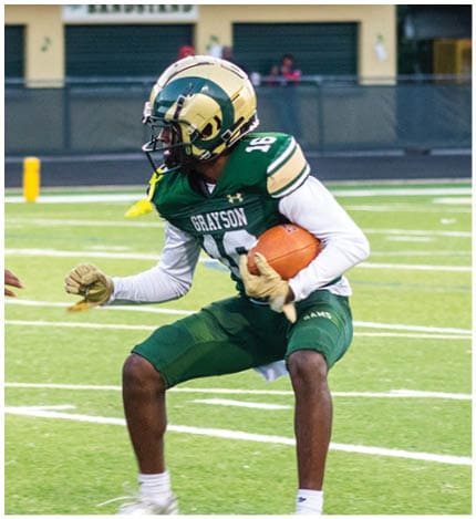 A person with long dreadlocks wearing a white football jersey with the name Grayson and the number 28 stands in front of two other people in similar football uniforms and helmets. Description generated by AI