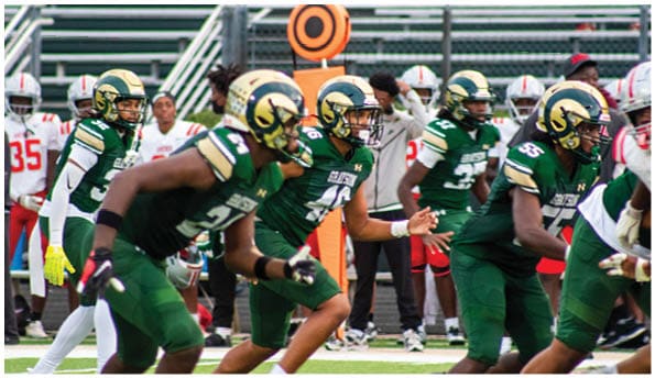 A group of football players in white uniforms and helmets run energetically through a torn banner held by cheerleaders on a field. Description generated by AI