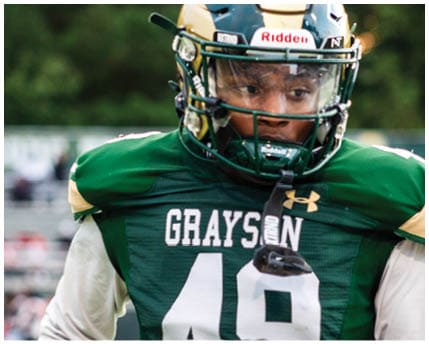 A person wearing a football helmet and a white jersey with the word Grayson and the number 10 stands outdoors with trees in the background. Description generated by AI