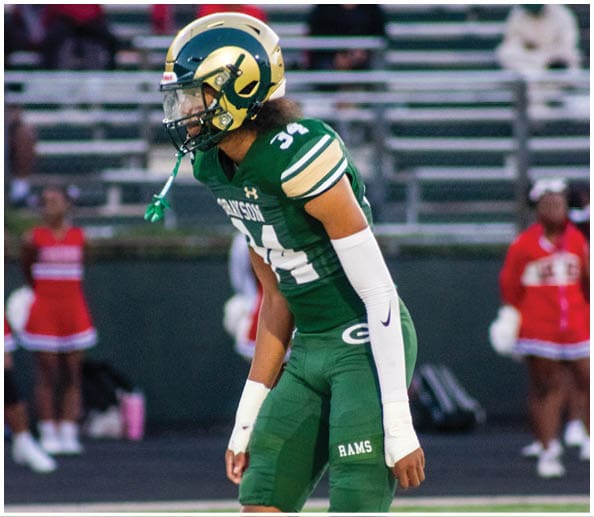 A person in a white and green football uniform with a gold helmet is on a field facing away from the camera while another person in a maroon and gold uniform stands in the background near empty bleachers. Description generated by AI