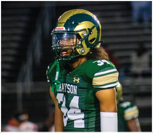 A person in a white and gold football uniform is sitting on the field holding a football while other players stand in the background. Description generated by AI