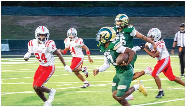 A person wearing a white football uniform and helmet is running with a football on a field while people in green cheerleading outfits stand in the background. Description generated by AI