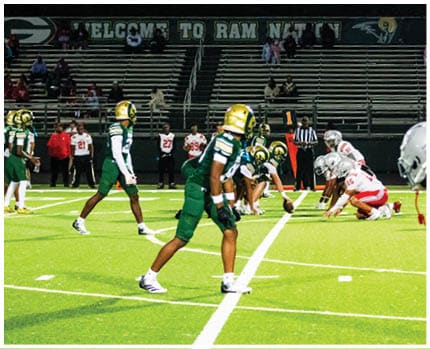 A person in a white and yellow football uniform is crouched on a green field with a referee and other players in the background. Description generated by AI