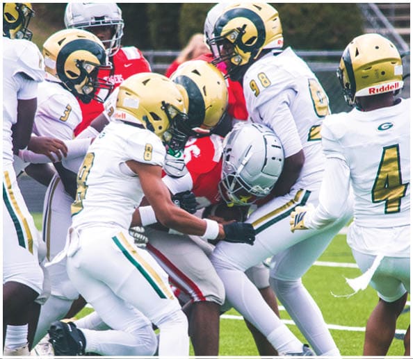 A person in a white and gold football uniform is crouched in an athletic stance on a green football field with other players and a coach in the background. Description generated by AI