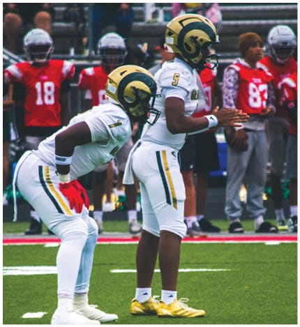 A person with long dreadlocks wearing a white football jersey with the name Grayson and the number 28 stands in front of two other people in similar football uniforms and helmets. Description generated by AI