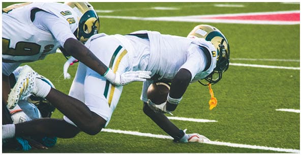 Two people wearing white sports uniforms with green and yellow stripes stand on a field with others in the background. Description generated by AI