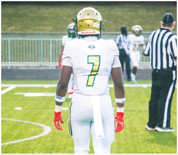 A person in a white and green football uniform with a gold helmet is on a field facing away from the camera while another person in a maroon and gold uniform stands in the background near empty bleachers. Description generated by AI