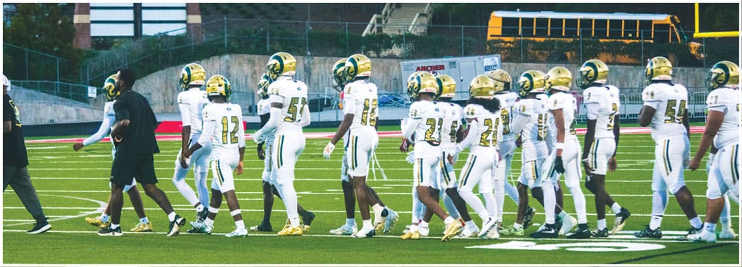 A person wearing a white sports uniform with the number 47 is standing on a field with other people in similar uniforms and spectators seated in bleachers in the background. Description generated by AI