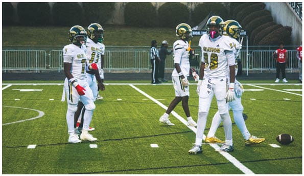 A person wearing a white football uniform and helmet is running with a football on a field while people in green cheerleading outfits stand in the background. Description generated by AI