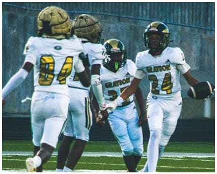 Three people wearing white and gold football uniforms and helmets are standing and interacting near a chain-link fence with trees and bleachers in the background. Description generated by AI