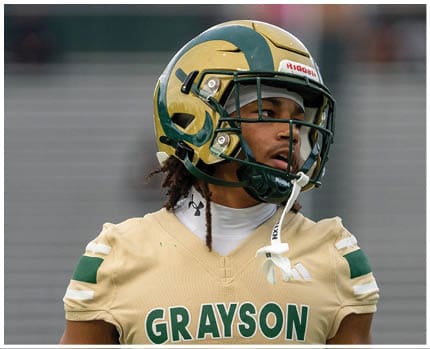 A person wearing a gold and black football helmet and a white jersey with the word “BRAVES" visible is looking to the side against a blue sky background. Description generated by AI