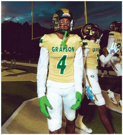 A person with long dreadlocks wearing a white football jersey with the name Grayson and the number 28 stands in front of two other people in similar football uniforms and helmets. Description generated by AI
