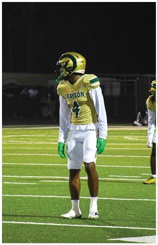 A person wearing white athletic pants and cleats is standing on a green football field with white yard lines. Description generated by AI