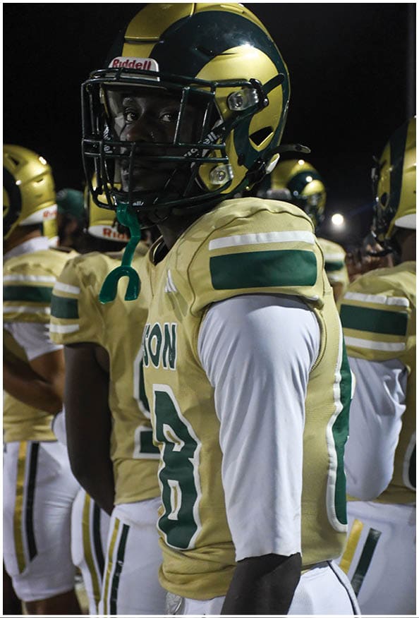 A person in a white and gold football uniform is crouched in an athletic stance on a green football field with other players and a coach in the background. Description generated by AI