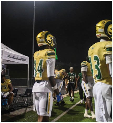 A person with long dreadlocks wearing a white football jersey with the name Grayson and the number 28 stands in front of two other people in similar football uniforms and helmets. Description generated by AI