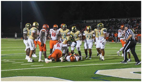 A group of football players in white uniforms and helmets run energetically through a torn banner held by cheerleaders on a field. Description generated by AI