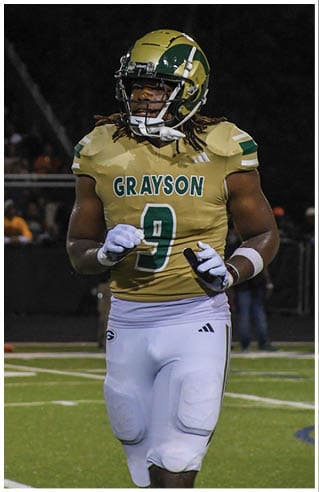 A person wearing white athletic pants and cleats is standing on a green football field with white yard lines. Description generated by AI