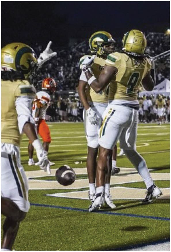 A person in a white and gold football uniform is crouched in an athletic stance on a green football field with other players and a coach in the background. Description generated by AI