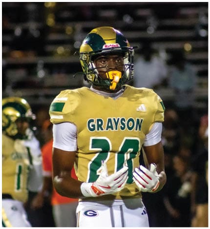 A person with long dreadlocks wearing a white football jersey with the name Grayson and the number 28 stands in front of two other people in similar football uniforms and helmets. Description generated by AI