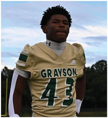 A person with long dreadlocks wearing a white football jersey with the name Grayson and the number 28 stands in front of two other people in similar football uniforms and helmets. Description generated by AI