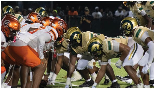 A group of football players in white uniforms and helmets run energetically through a torn banner held by cheerleaders on a field. Description generated by AI