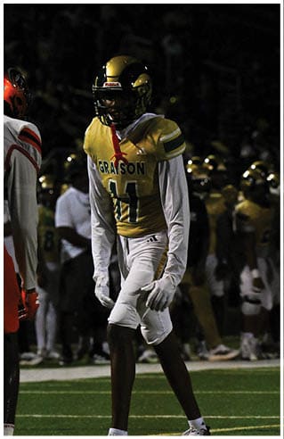 A person wearing white athletic pants and cleats is standing on a green football field with white yard lines. Description generated by AI
