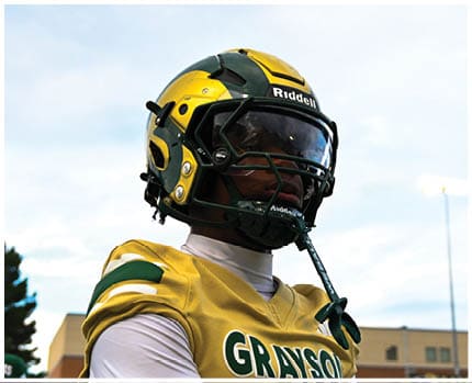A person wearing a gold and black football helmet and a white jersey with the word “BRAVES" visible is looking to the side against a blue sky background. Description generated by AI
