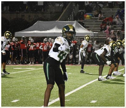 Two people in white and green football uniforms stand on a field with one clenching a fist and a referee and opposing players in red uniforms visible in the background. Description generated by AI