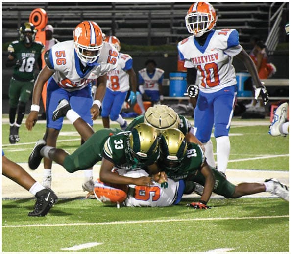 A person in a white and gold football uniform is crouched in an athletic stance on a green football field with other players and a coach in the background. Description generated by AI