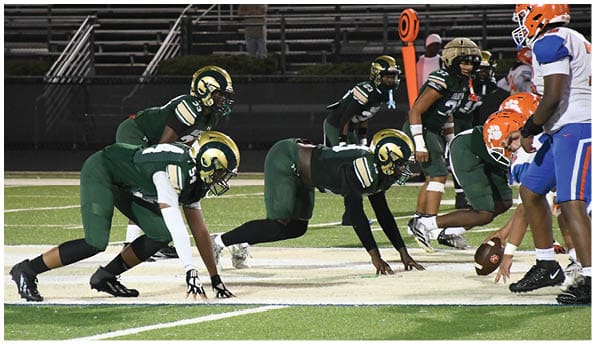 A group of football players in white uniforms and helmets run energetically through a torn banner held by cheerleaders on a field. Description generated by AI