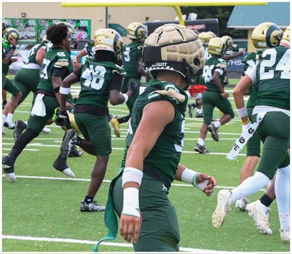 A person in a white and green football uniform with a gold helmet is on a field facing away from the camera while another person in a maroon and gold uniform stands in the background near empty bleachers. Description generated by AI