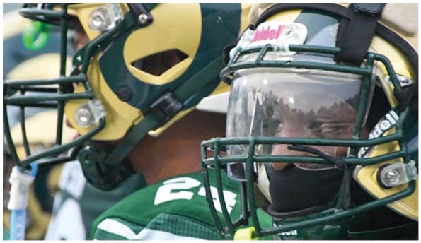 A person wearing a white football uniform and helmet is running with a football on a field while people in green cheerleading outfits stand in the background. Description generated by AI
