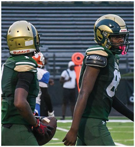 A person with long dreadlocks wearing a white football jersey with the name Grayson and the number 28 stands in front of two other people in similar football uniforms and helmets. Description generated by AI