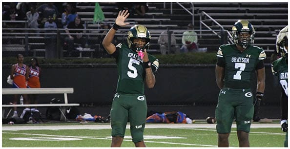 Two people wearing white sports uniforms with green and yellow stripes stand on a field with others in the background. Description generated by AI