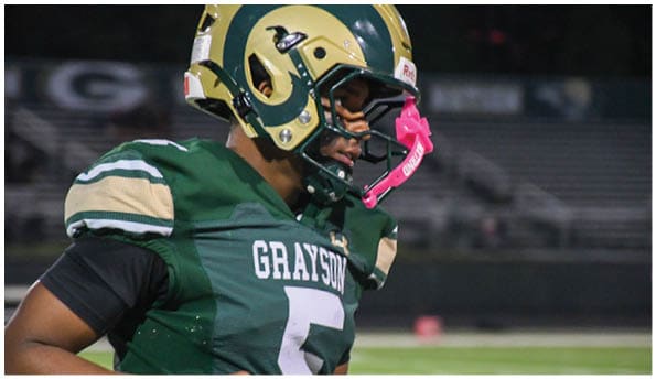 A person wearing a white football uniform and helmet is running with a football on a field while people in green cheerleading outfits stand in the background. Description generated by AI
